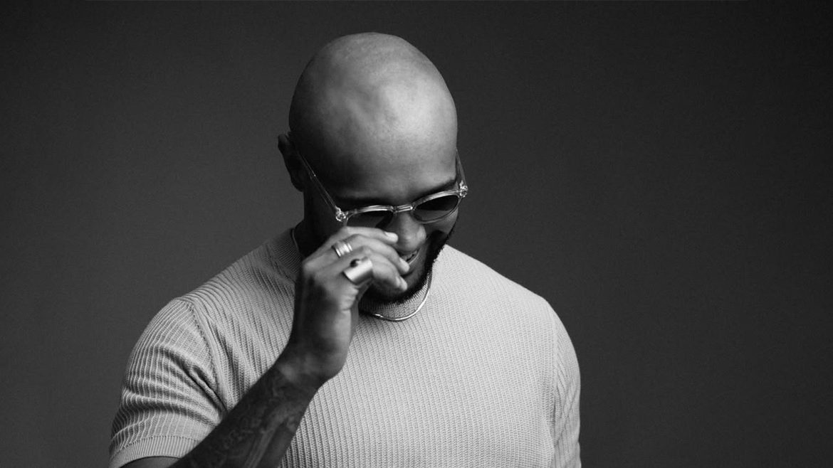 El Mukuka in a black-and-white studio shot, captured mid-thought with headphones around his neck, exuding creative focus.